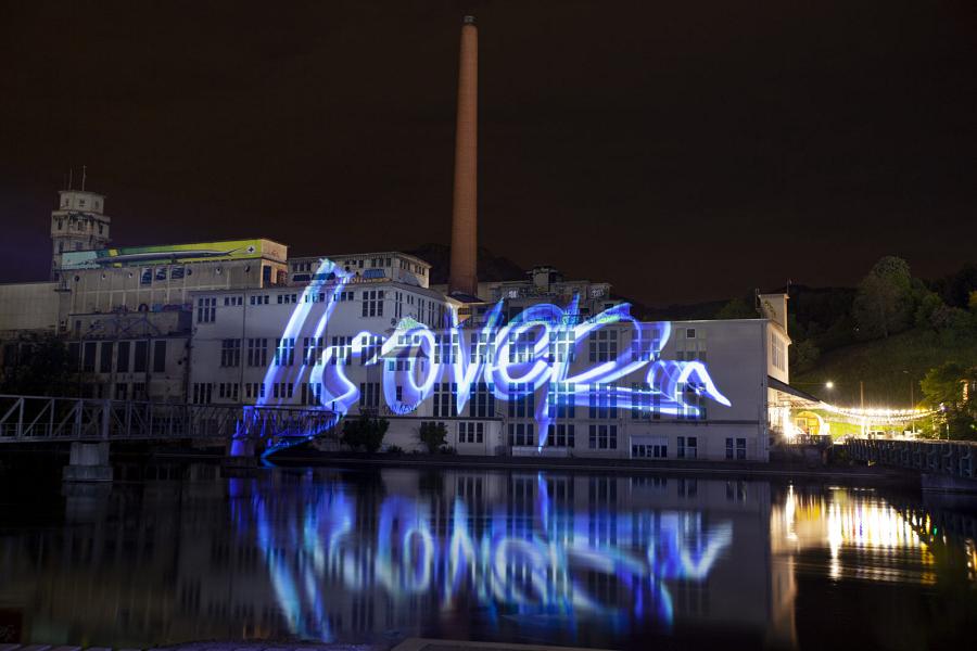 Fabrikgebäude mit Schornstein bei Nacht, blaue Lichtschrift auf der Fassade und im Wasser gespiegelt
