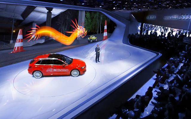 Rotes Auto auf einer weißen Bühne vor Publikum, dahinter eine orangefarbene Lichtzeichnung