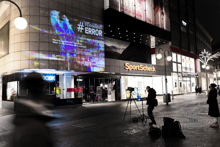 Einkaufsstraße bei Nacht, an einer Fassade eine Projektion mit dem Hashtag „#Permanent Error“, davor ein Fotograf mit Stativ