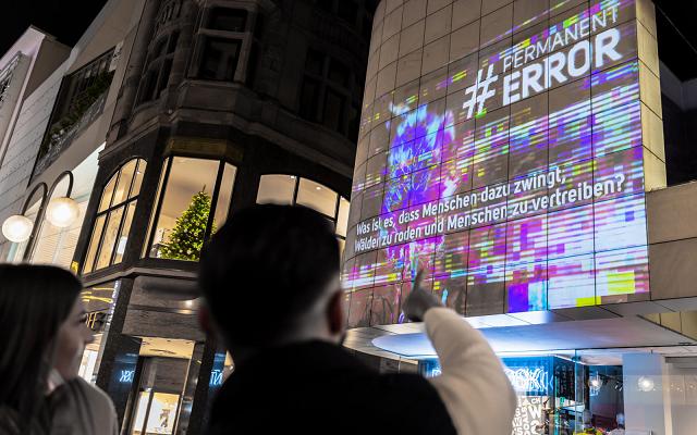 Zwei Personen zeigen nachts auf eine Projektion mit dem Schriftzug „#Permanent Error“ auf einer Hausfassade