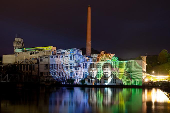 Alte Fabrikanlage mit Schornstein bei Nacht, bunte Lichtprojektion mit Gesichtern auf der Fassade, gespiegelt im Wasser