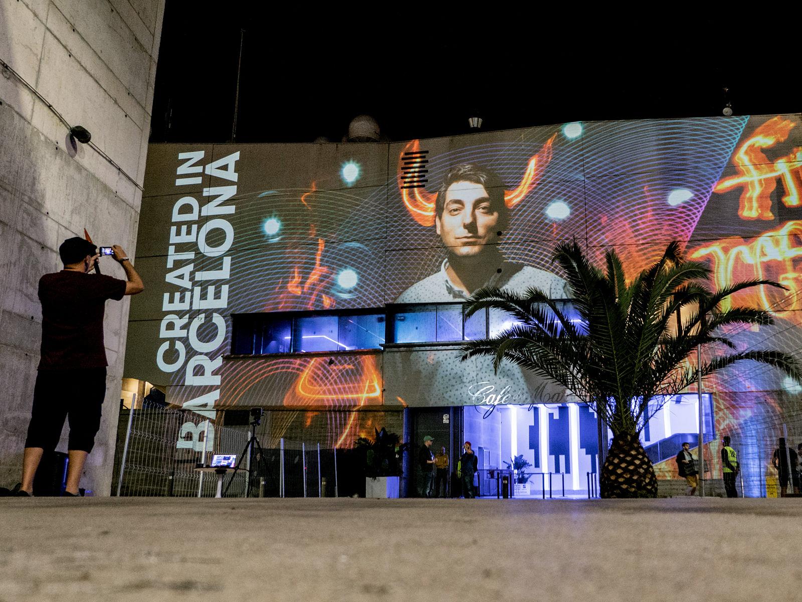 Gebäudefassade mit Projektion eines Gesichts und dem Schriftzug „Created in Barcelona“, davor eine Palme