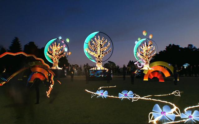 Parkfläche in der Dämmerung mit leuchtenden Baumskulpturen und Lichtblumen am Boden