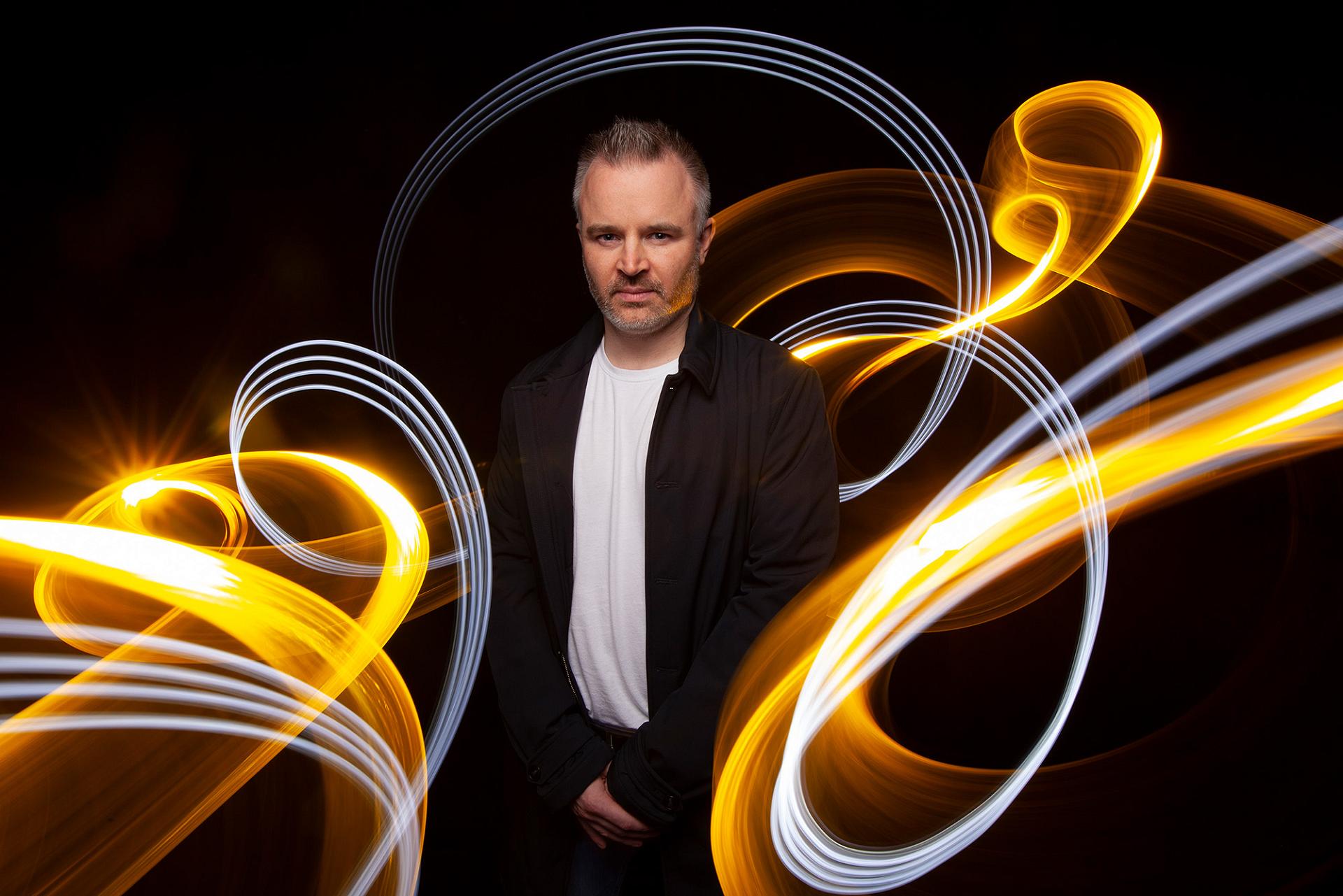 Man in a black jacket standing against a dark background between golden-yellow and white light trails
