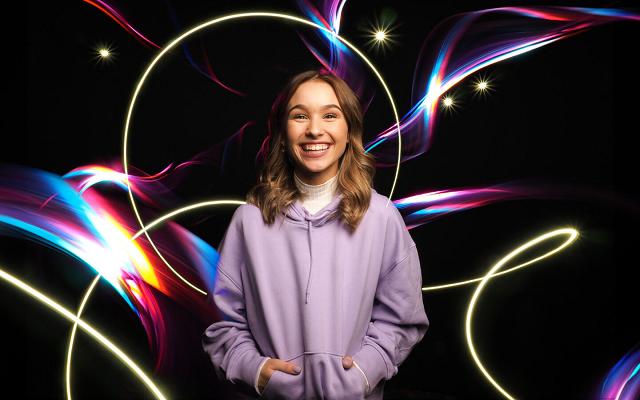 Lächelnde junge Frau im lila Hoodie vor schwarzem Hintergrund, umgeben von bunten Lichtspuren und Lichtkreisen