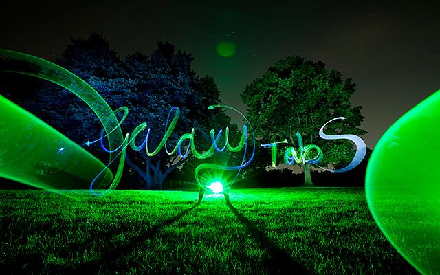 Green light drawing with the words “Galaxy Tab S” on a meadow in front of trees at night