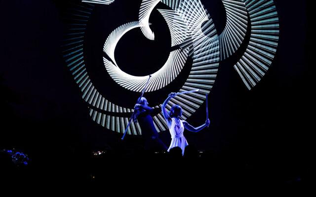 Zwei Performer auf einer dunklen Bühne vor einer großen Spirale aus weißen Lichtlinien
