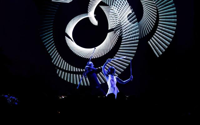 Zwei Performer auf einer dunklen Bühne vor einer großen Spirale aus weißen Lichtlinien