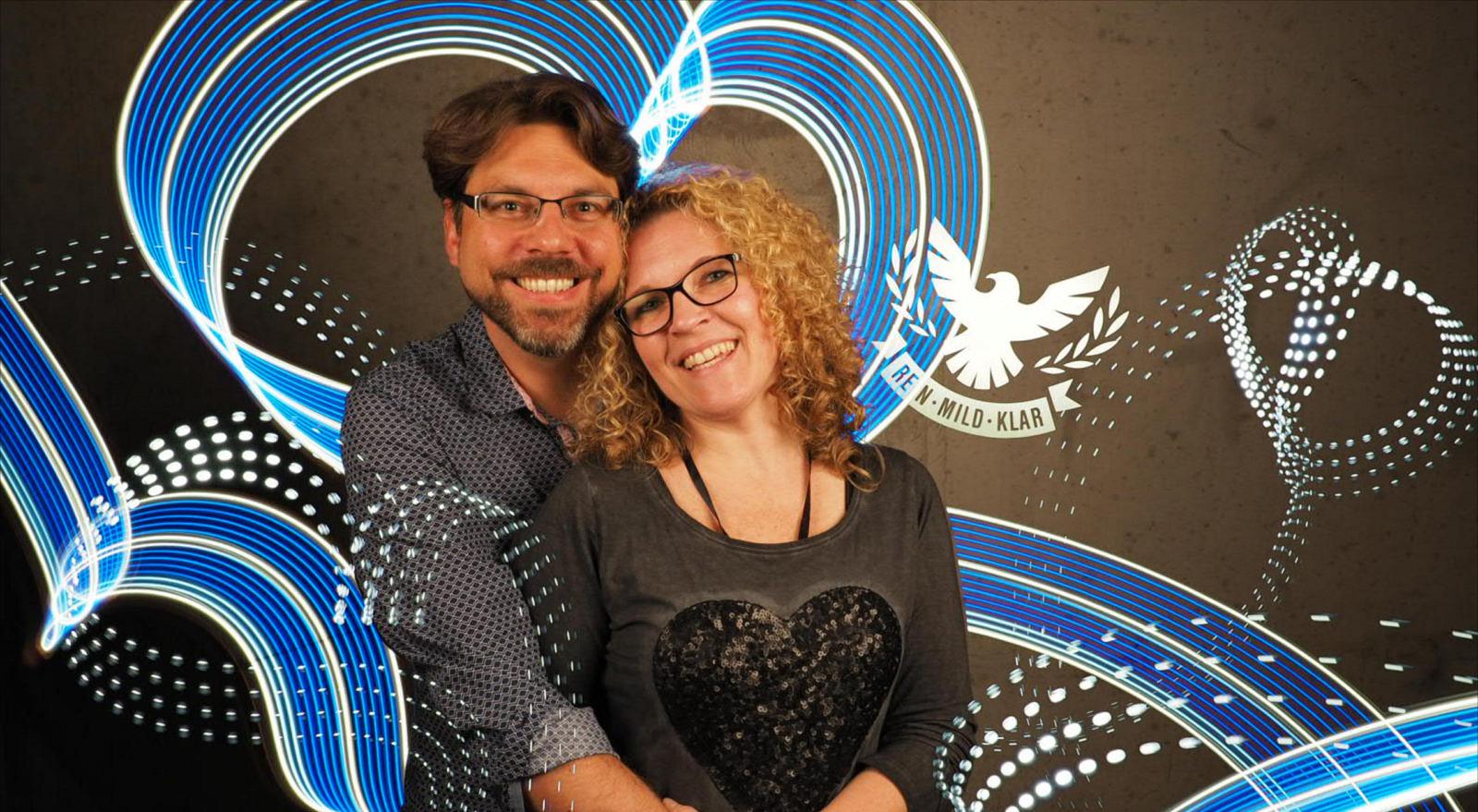 Paar vor blauen Lichtschleifen in Herzform, rechts ein Adler-Logo