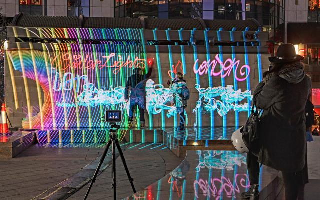 Passanten und Kamera auf Stativ vor einer Wand mit bunter Lichtprojektion und Light-Graffiti