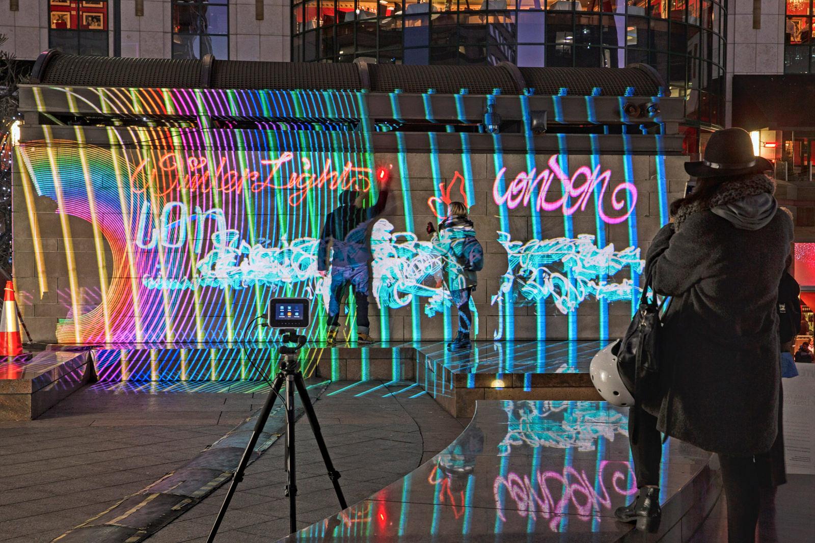 Passanten und Kamera auf Stativ vor einer Wand mit bunter Lichtprojektion und Light-Graffiti