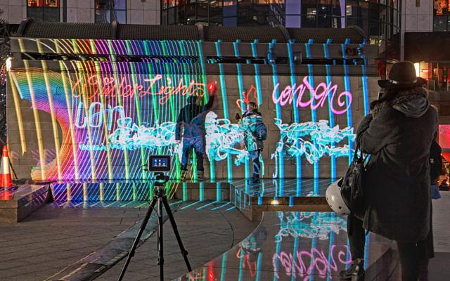 Passanten und Kamera auf Stativ vor einer Wand mit bunter Lichtprojektion und Light-Graffiti