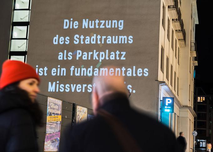 Passanten vor einer Hauswand mit projiziertem Text zur Nutzung des Stadtraums als Parkplatz
