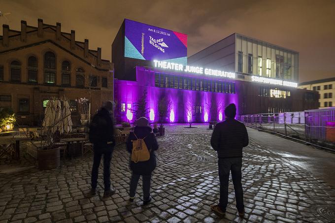 Theatergebäude in violettem Licht mit Projektion zum Tag der Bildung, davor drei Personen