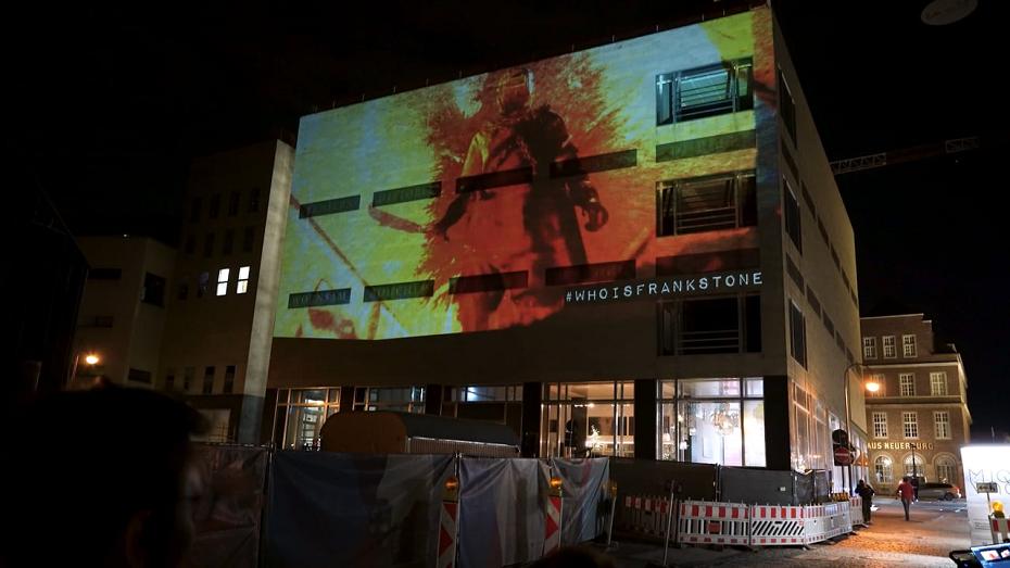 Gebäude bei Nacht mit großer Projektion in Rot und Gelb auf der Fassade, darunter der Hashtag „#WhoIsFrankStone“