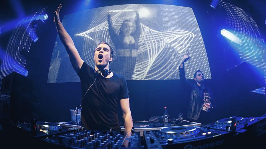 DJ mit erhobenem Arm am Mischpult vor einer LED-Wand mit Lichtmustern in einem Club