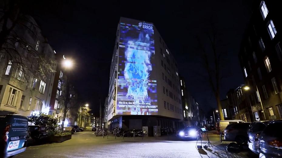Hohes Wohnhaus bei Nacht, auf die Fassade ist eine blaue Projektion mit dem Hashtag „#Error“ geworfen